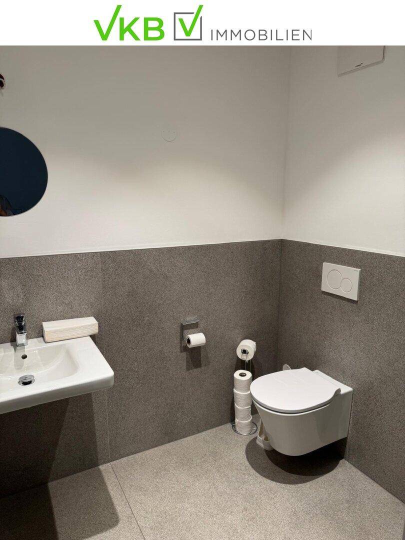 Modernes Gäste-WC mit wandhängender Toilette, kleinem Waschbecken und grauen Fliesen, minimalistisch gestaltet.
