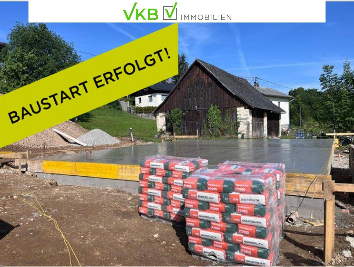 Baustelle mit frisch gegossener Betonplatte und Baumaterialien unter blauem Himmel, daneben ein altes Holzhaus.