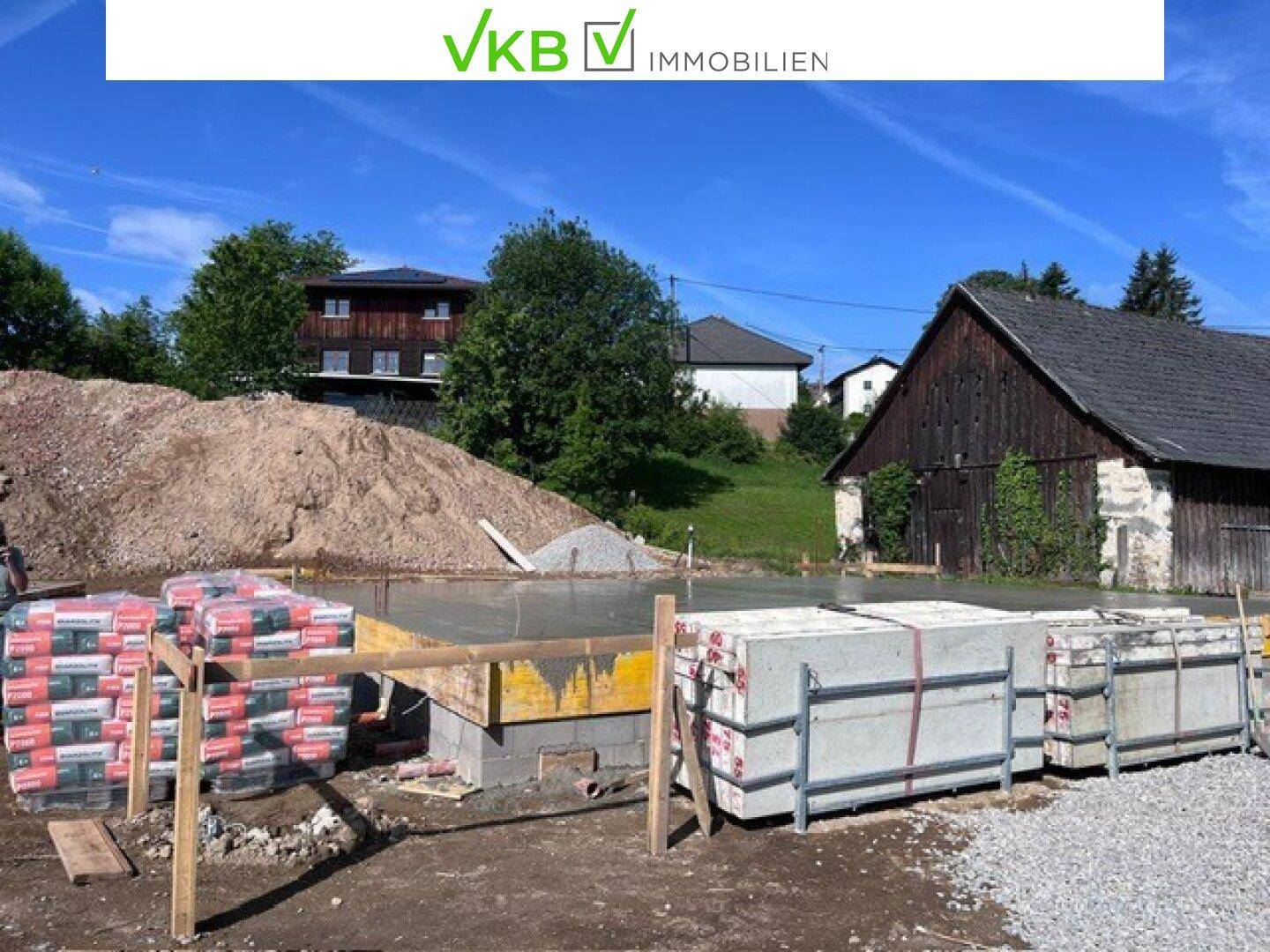 Baustelle mit vorbereitetem Fundament und Baumaterialien, umgeben von ländlicher Bebauung und viel Grün.