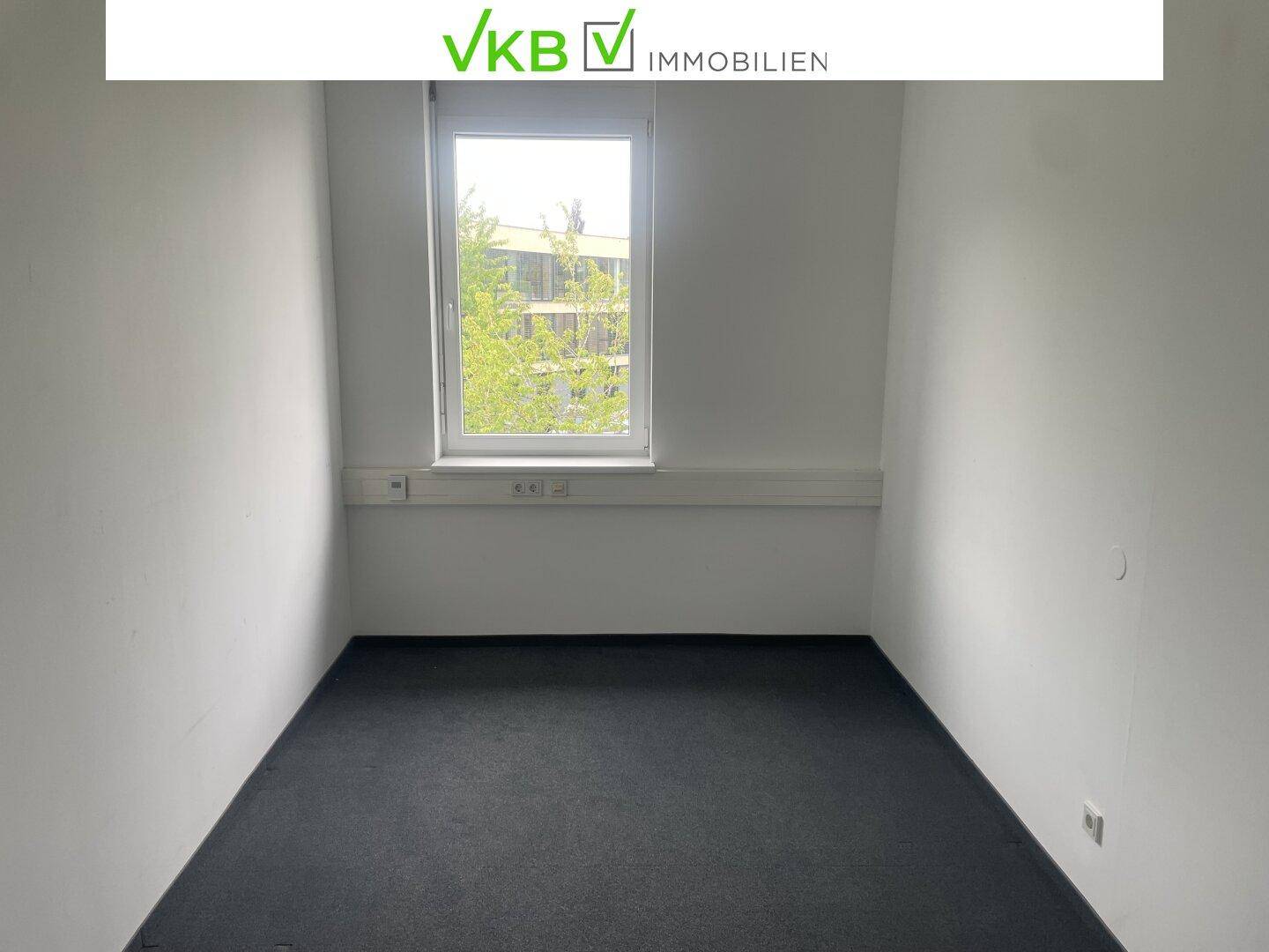 Kleines, leeres Büro mit einem Fenster, dunklem Teppichboden und weißen Wänden.