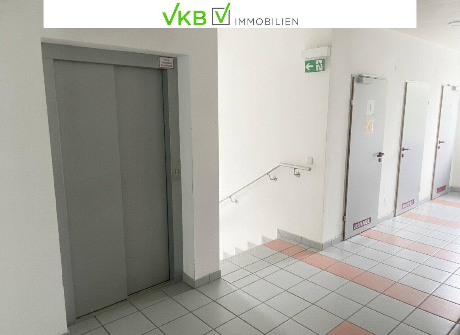 Flur mit Aufzug, Treppenhaus und mehreren Türen, ausgestattet mit Fliesenboden und Handlauf.