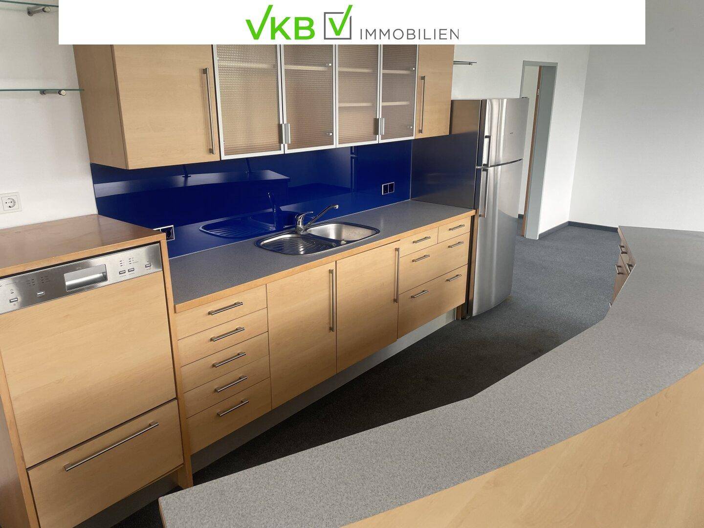 Detailansicht der Teeküche mit Spülmaschine, Spüle und Holzschränken unter blauer Rückwand.