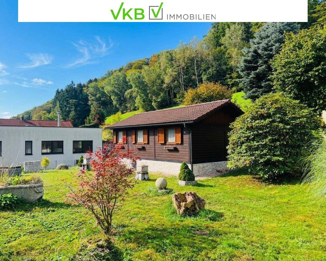 Gartenlandschaft mit einem charmanten Holzhaus und einem modernen Gebäude im Hintergrund unter blauem Himmel.