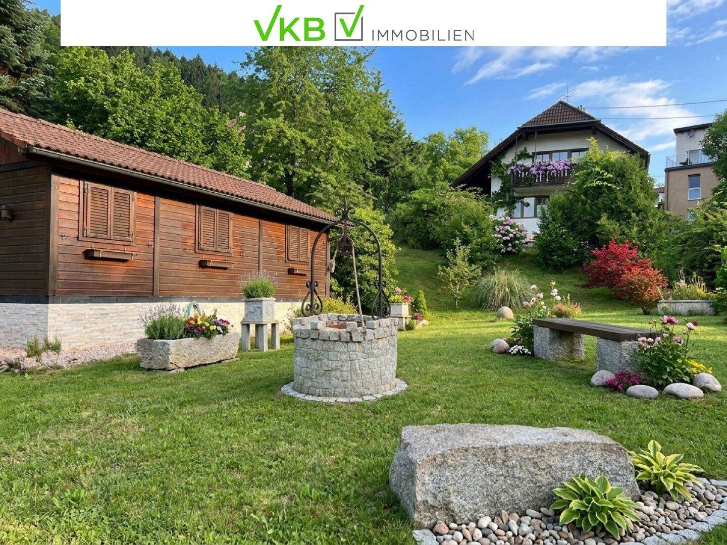 Weitläufiger Garten mit einem traditionellen Brunnen, Sitzbank und dem rustikalen Holzhaus im Hintergrund.