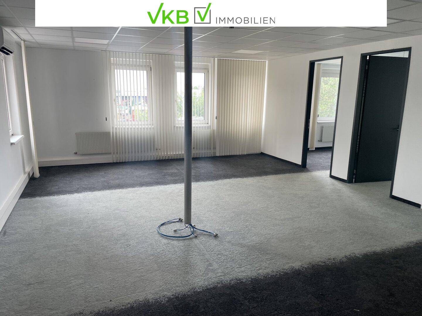 Geräumiges Büro mit Teppichboden in zwei Grautönen und vertikalen Jalousien an den Fenstern.