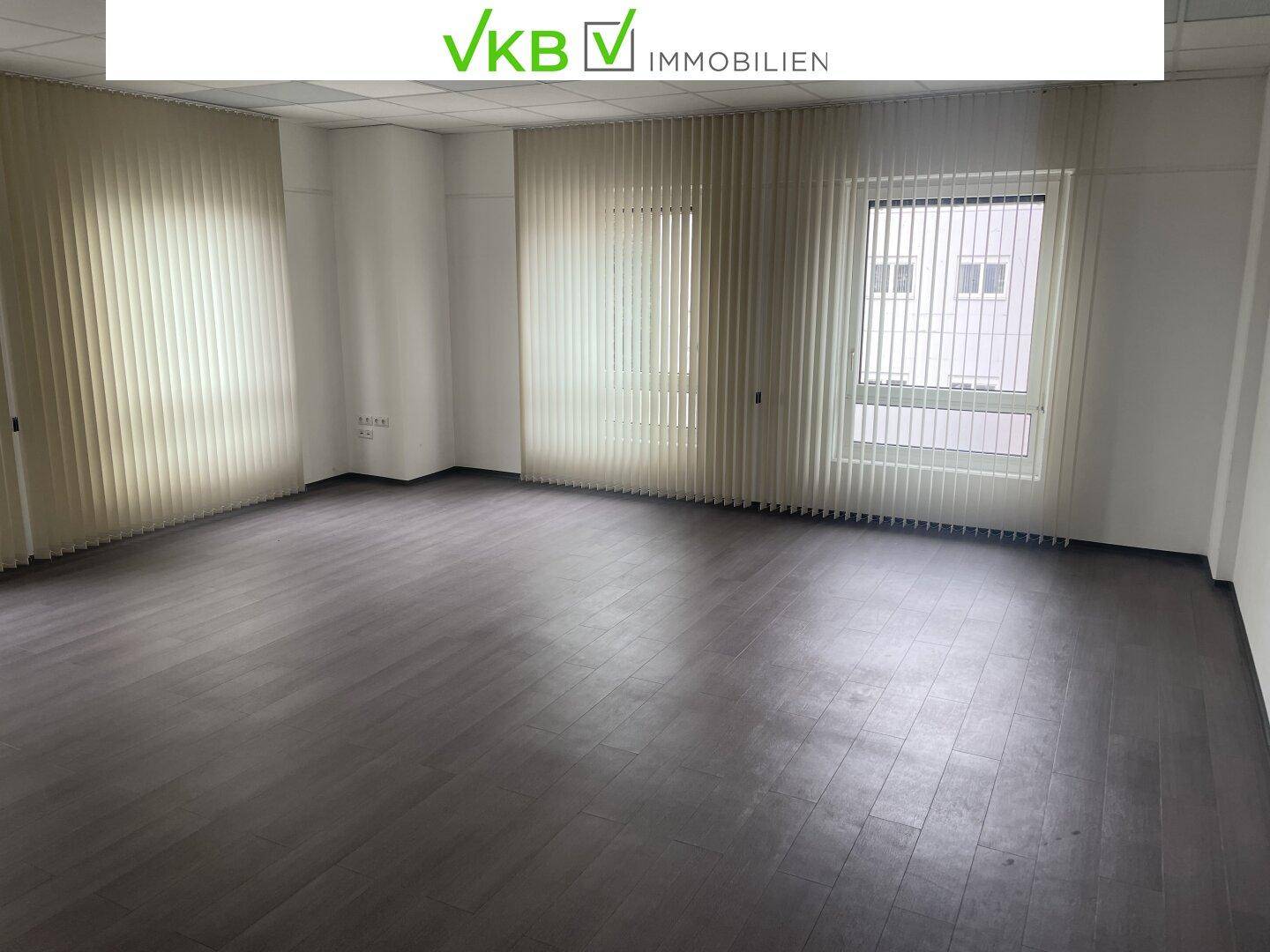 Großes Büro mit dunklem Laminatboden und vertikalen Jalousien an den Fenstern.