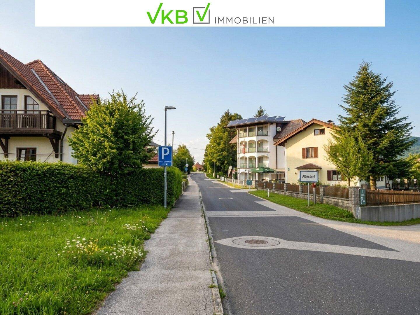 Ruhige Dorfstraße mit Wohnhäusern, grünen Hecken und einem Parkplatzschild unter klarem Himmel.