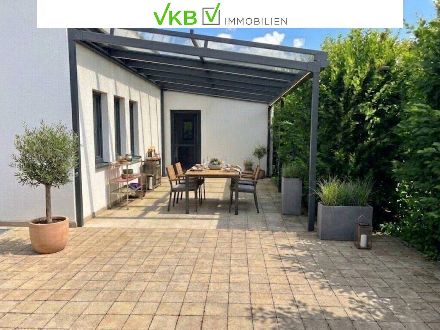 Überdachte Terrasse mit Esstisch und Stühlen, umgeben von Pflanzen und einem Olivenbaum.
