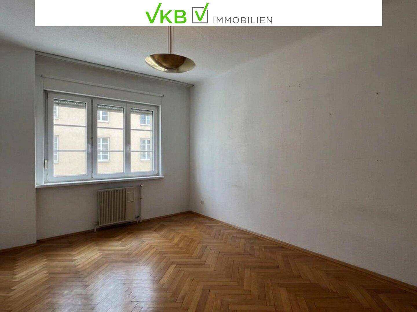 Helles Zimmer mit Fischgrätparkettboden, großem Fenster und Deckenleuchte.
