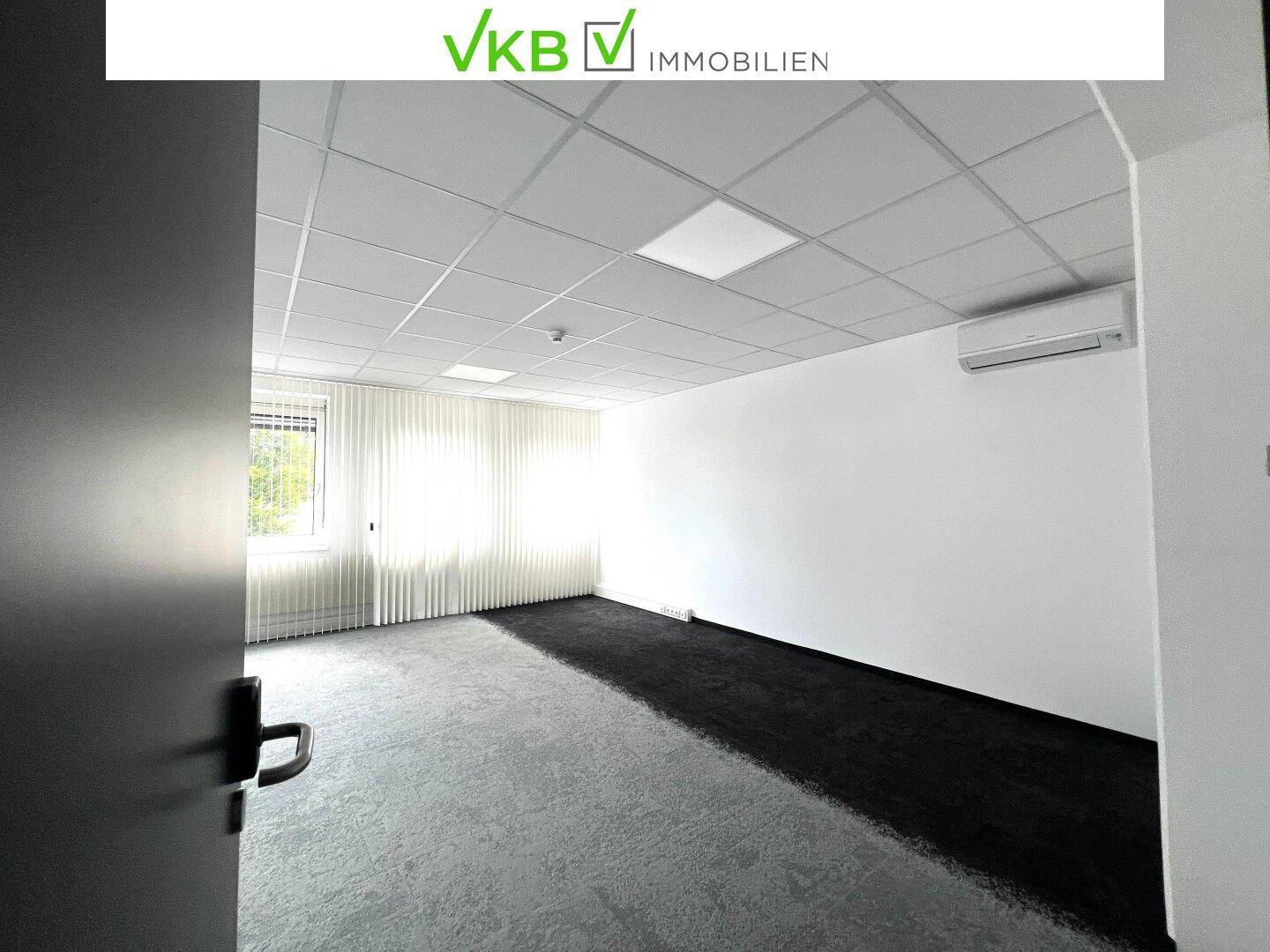 Leeres Büro mit Teppichboden, Klimaanlage und großen Fenstern, die viel Tageslicht hereinlassen.