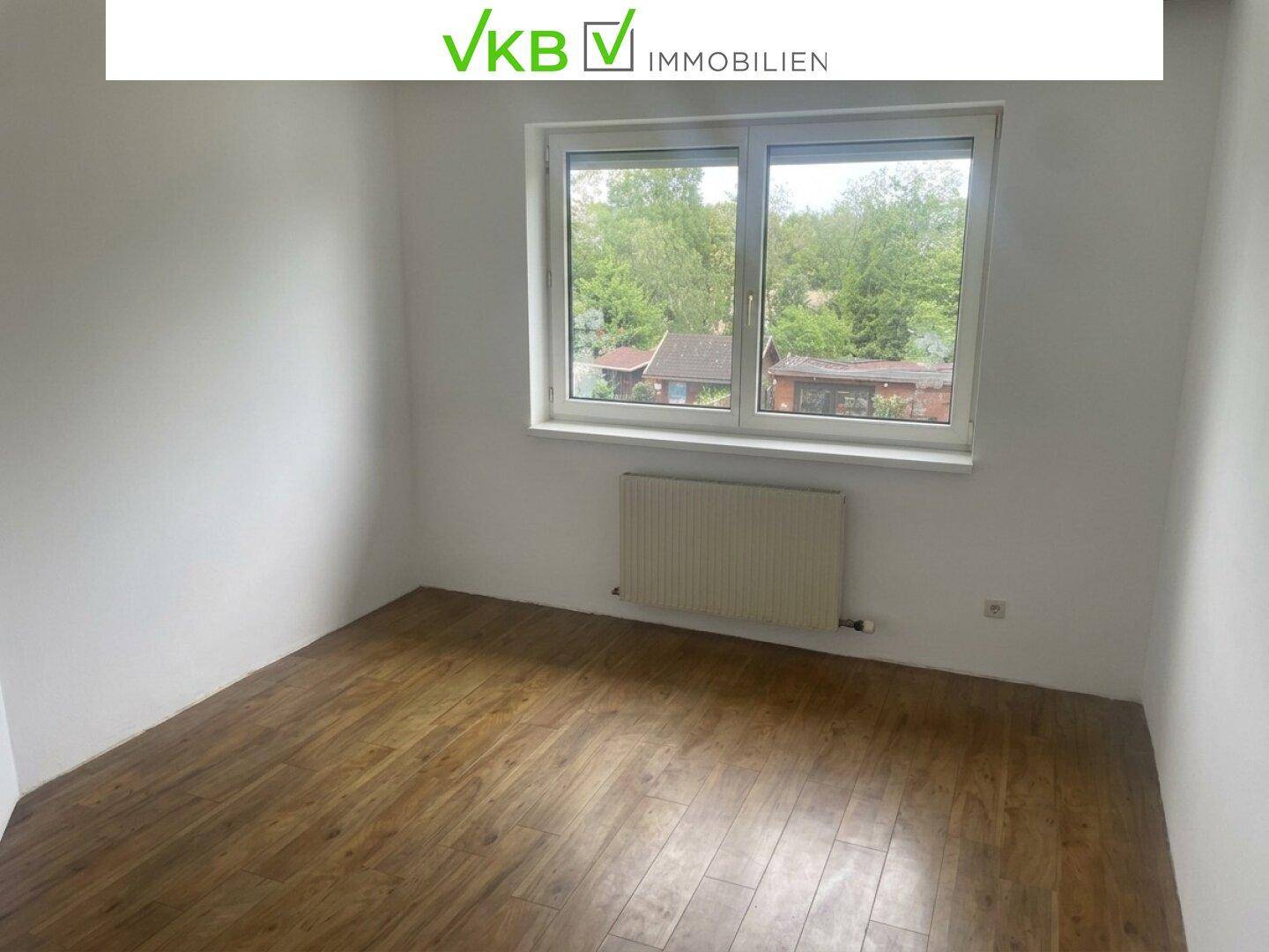 Helles Zimmer mit Laminatboden, Heizkörper und Fenster mit Blick ins Grüne.