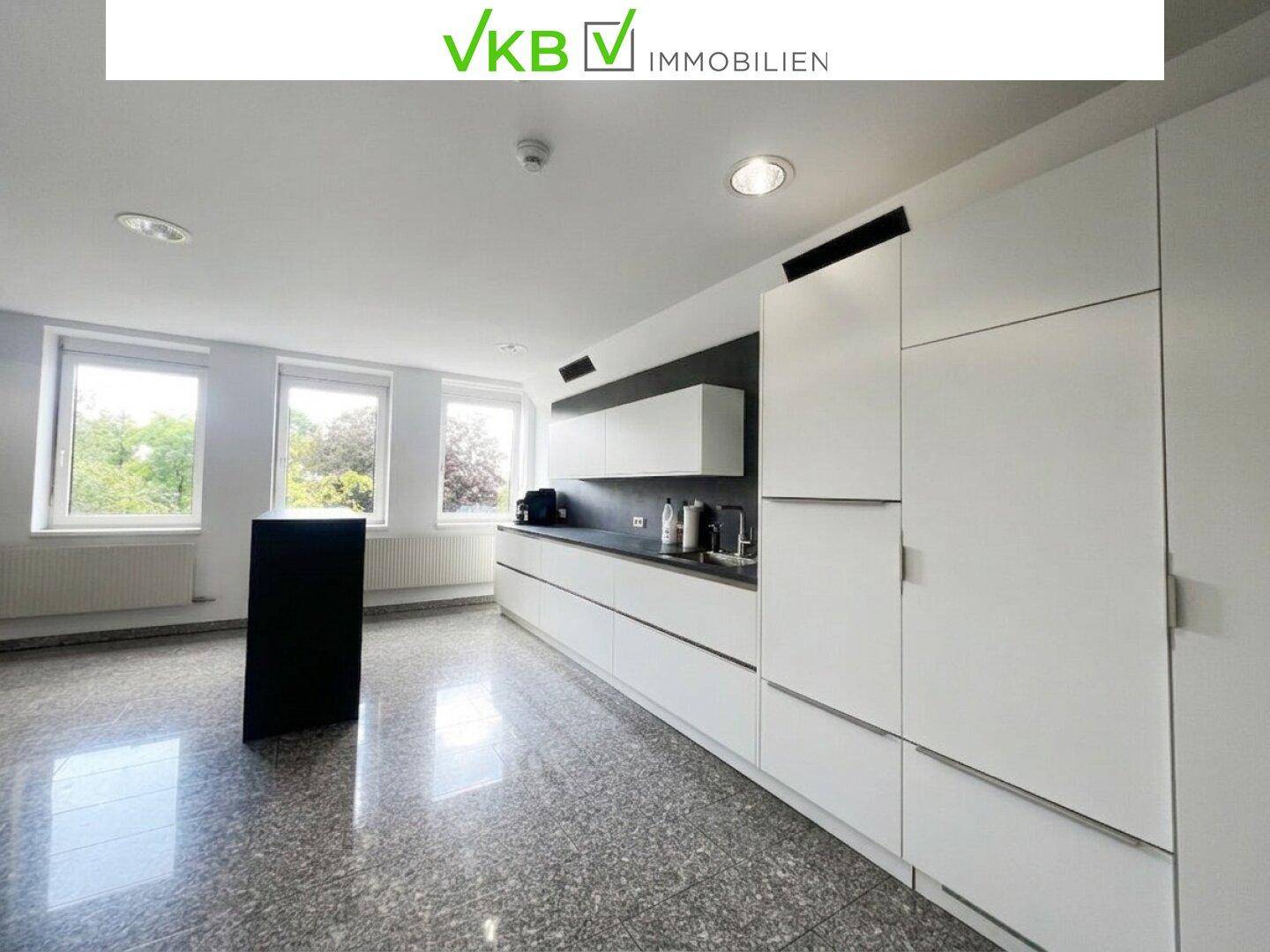 Helle, moderne Küche mit weißen Einbauschränken, dunkler Arbeitsplatte und großen Fenstern mit Blick ins Grüne.