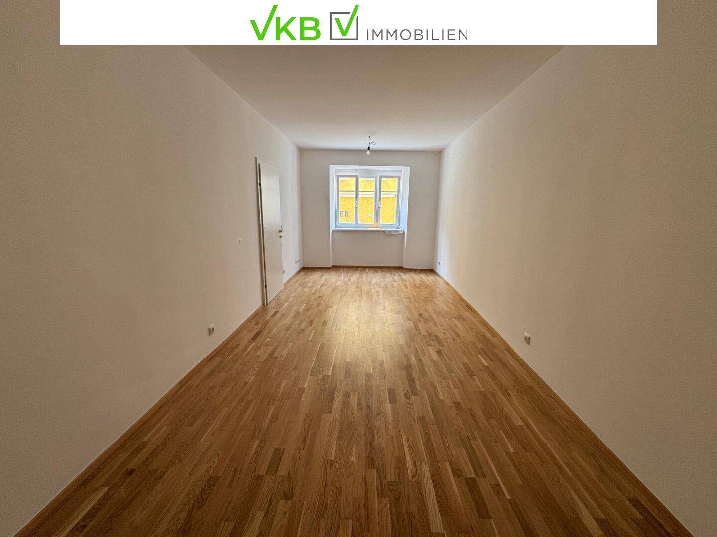 Geräumiges Wohnzimmer mit Holzboden und einem Fenster, das viel Tageslicht hereinlässt.