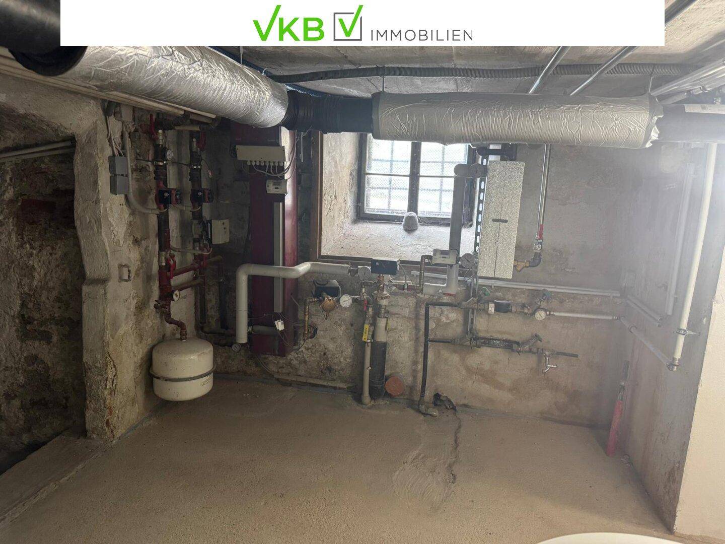Heizungsraum im Keller mit sichtbaren Rohren und technischen Installationen sowie einem kleinen Fenster.