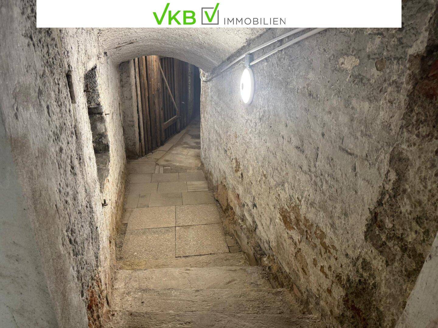 Historischer Kellergang mit Steinmauern und gewölbter Decke, der zu weiteren Räumen führt.
