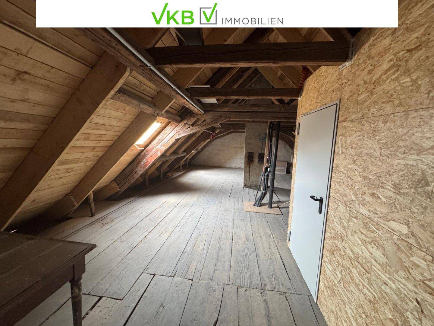 Weiterer Blick in den Dachboden mit Holzbalken, Dachschrägen und einer einfachen Tür, viel Potenzial.