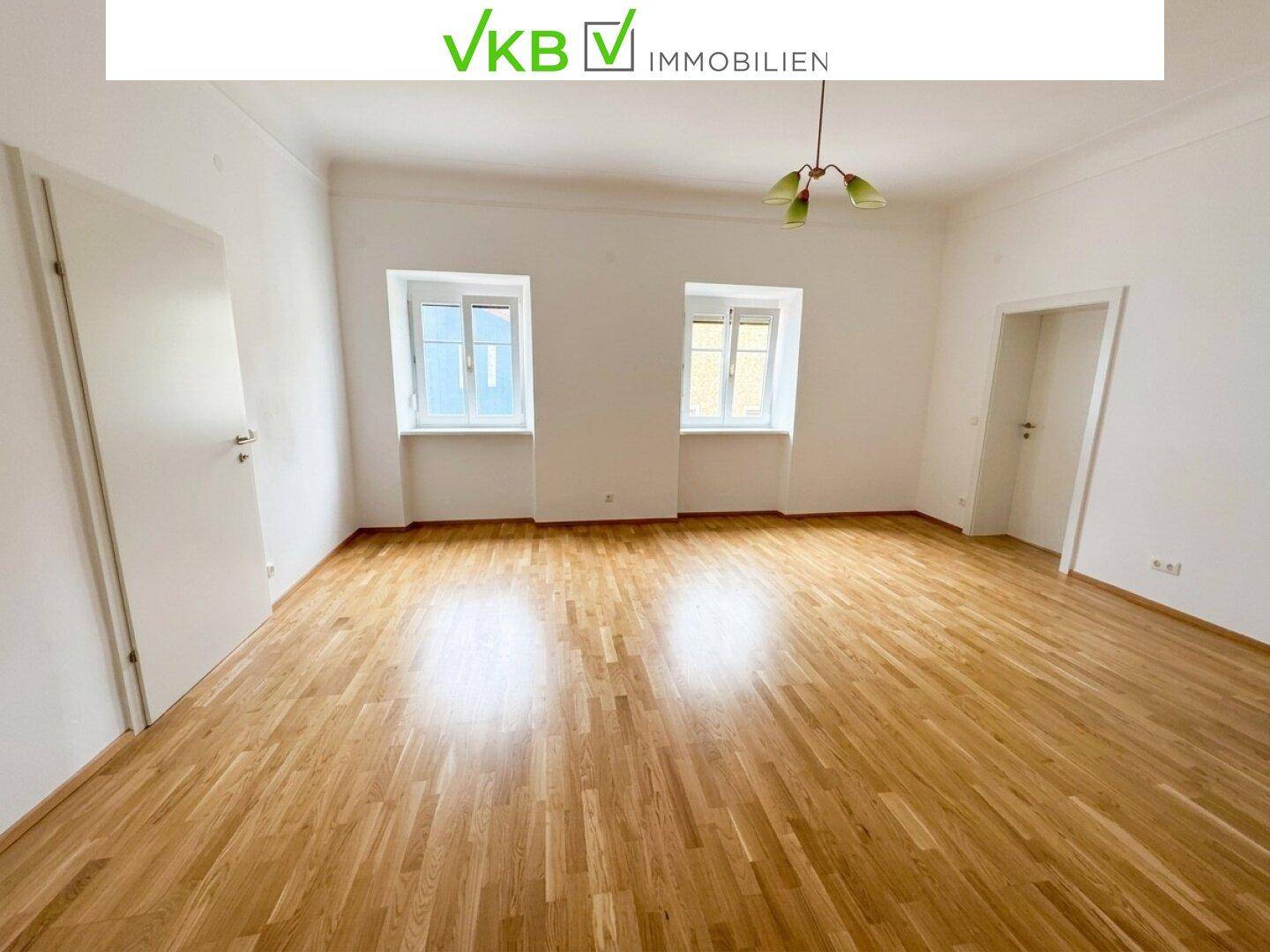 Großes Wohnzimmer mit Holzboden und zwei Fenstern, die für eine helle Atmosphäre sorgen.