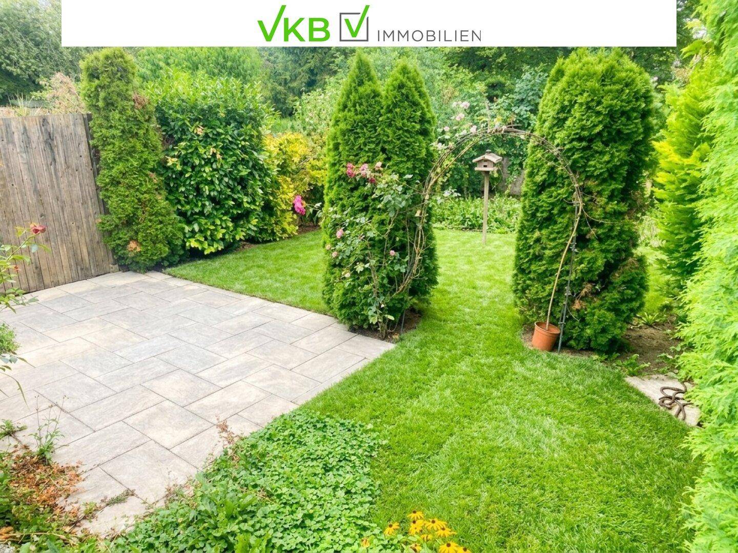 Gepflegter Garten mit Rasenfläche, Steinplattenweg und hohen Nadelbäumen.