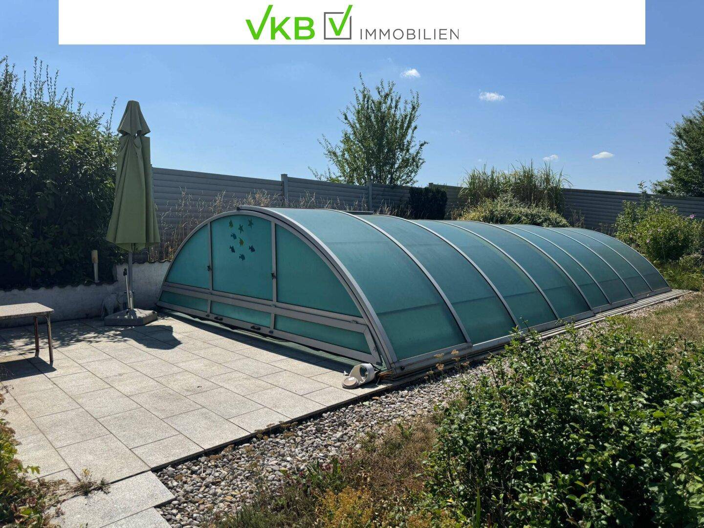 Überdachter Außenpool mit gepflasterter Terrasse und Sonnenschirm, umgeben von grüner Bepflanzung.