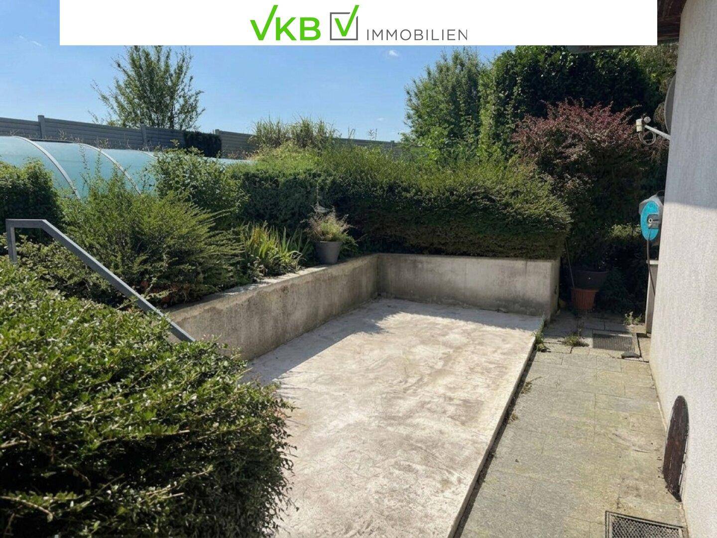 Betonierte Außenfläche, umgeben von hohen Hecken und einer Stützmauer, ideal für eine Terrasse.