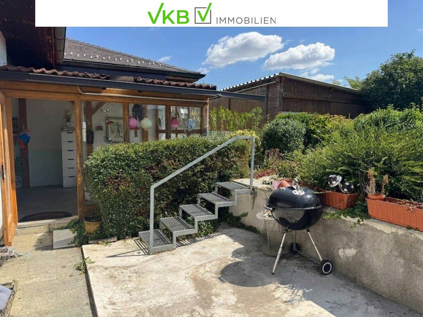 Einfacher Terrassenbereich mit Grill und Zugang zum Wintergarten, umgeben von Hecken und Pflanzen.
