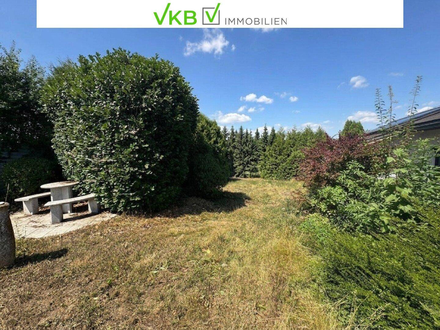 Großer Gartenbereich mit Steintisch und Bänken, umgeben von üppigen Bäumen und Sträuchern.