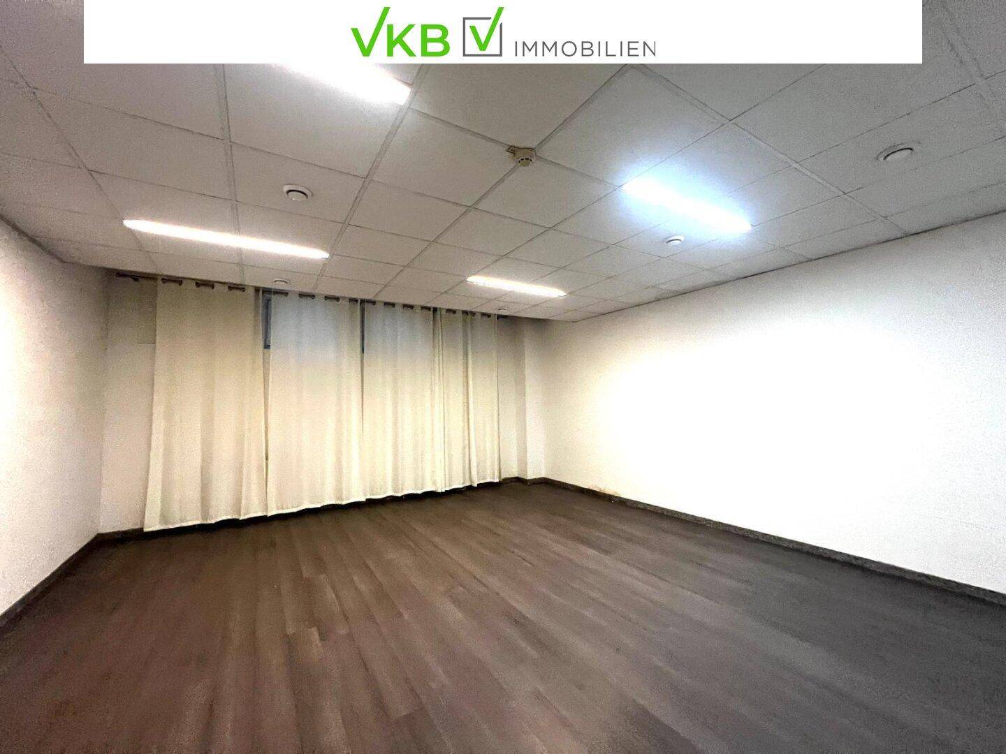 Heller Büroraum mit dunklem Holzboden und Vorhängen, ausgestattet mit Rasterdeckenbeleuchtung, bereit zur Nutzung.