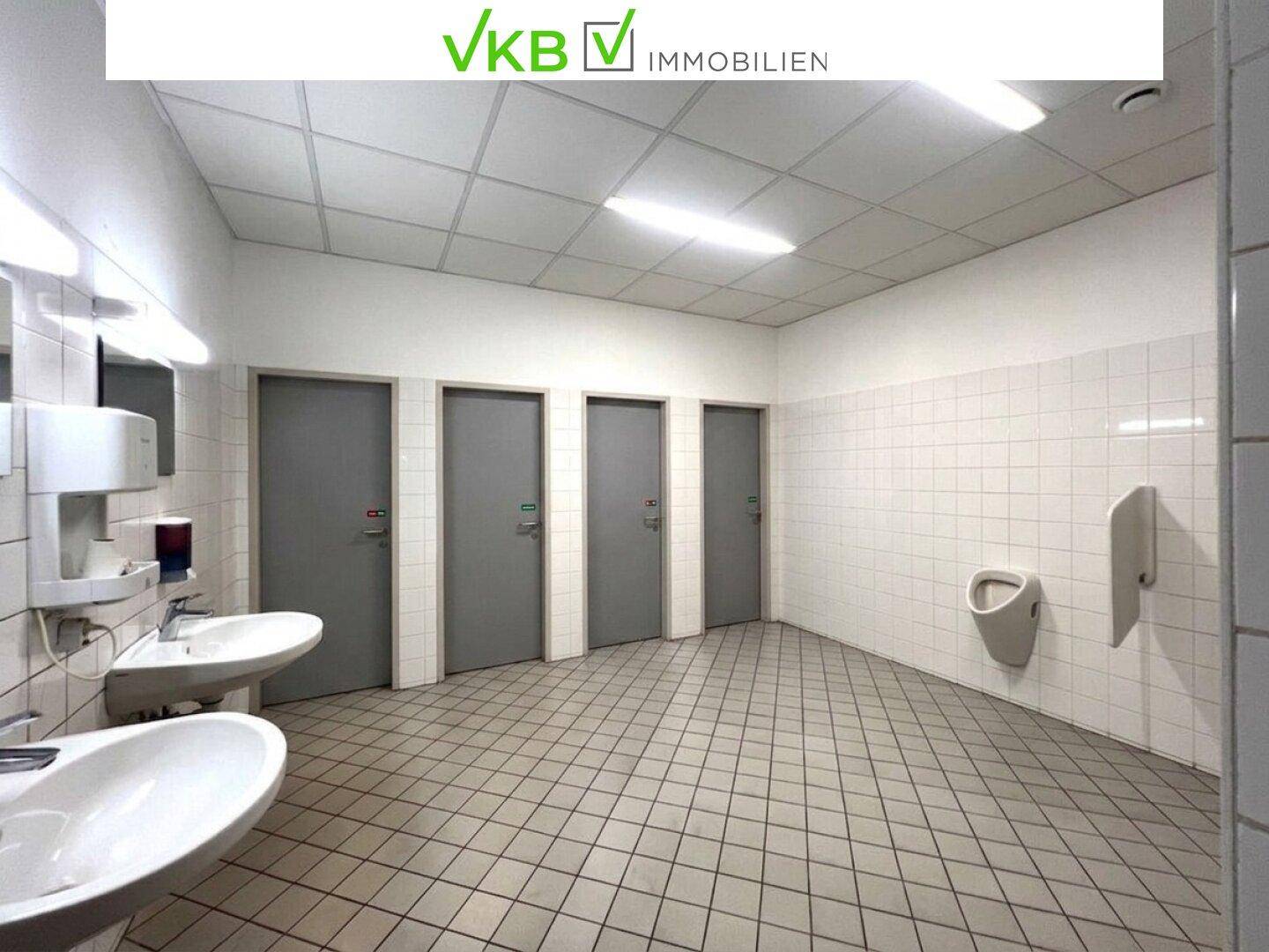 Sanitärbereich mit mehreren Waschbecken, Toilettenkabinen und einem Urinal, gefliest und funktional ausgestattet.