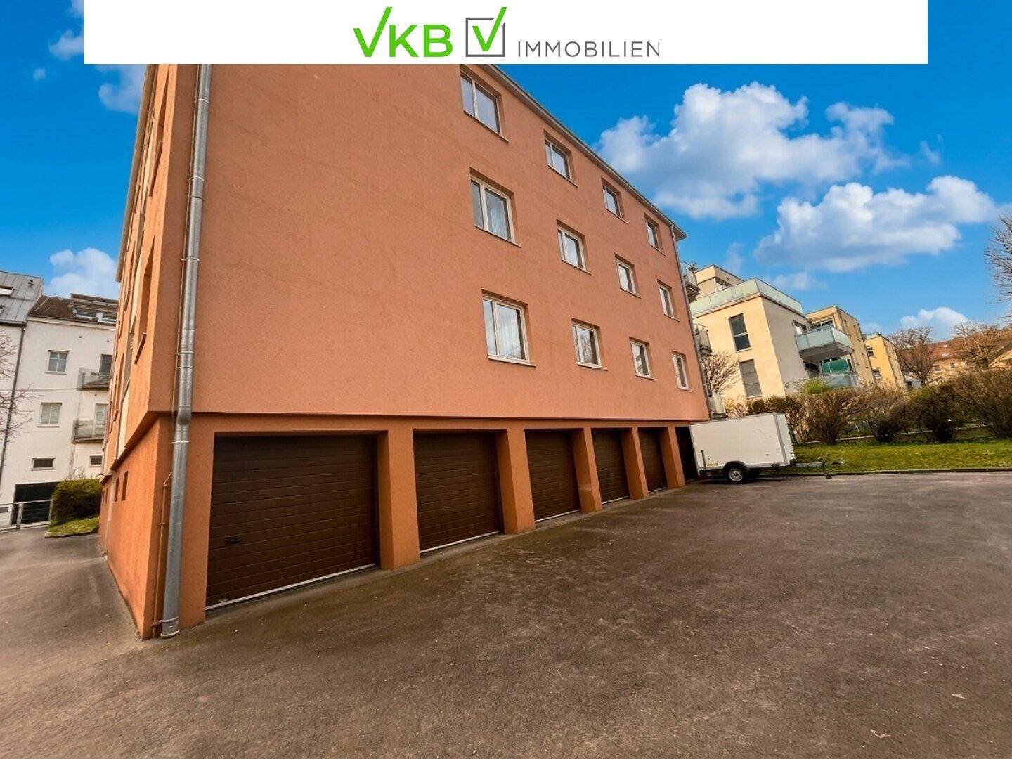 Seitenansicht des Gebäudes mit mehreren Garagentoren im Erdgeschoss und einem gepflasterten Vorplatz.