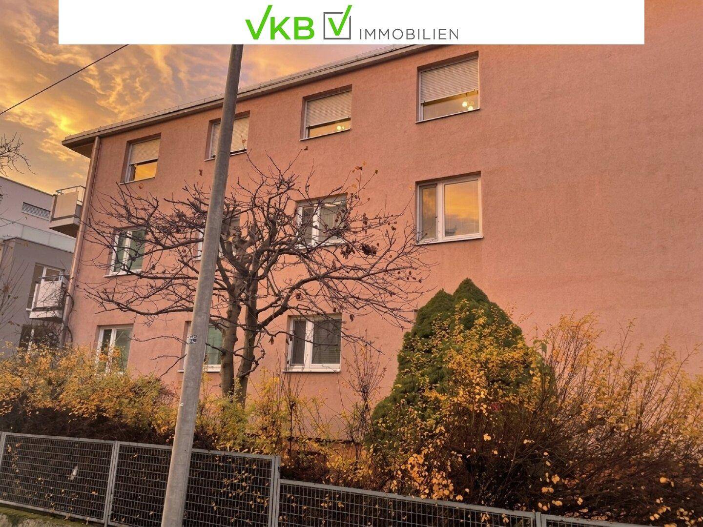 Gebäudeansicht mit herbstlichen Bäumen und Sträuchern vor der Fassade unter einem dramatischen Himmel.