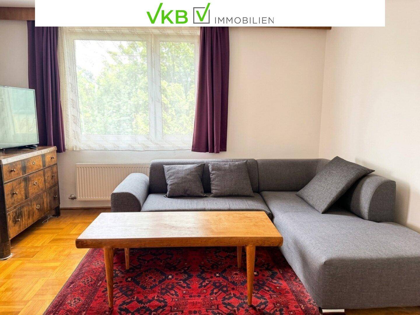 Heller Wohnbereich mit einem grauen Ecksofa, Holztisch und einem roten Teppich vor dem Fenster.