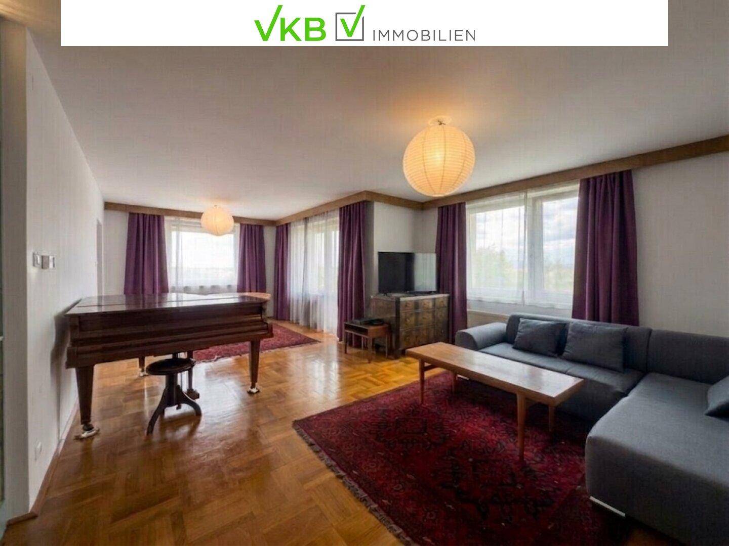 Geräumiges Wohnzimmer mit Parkettboden, einem Flügel und einem großen Sofa unter zwei Hängelampen.