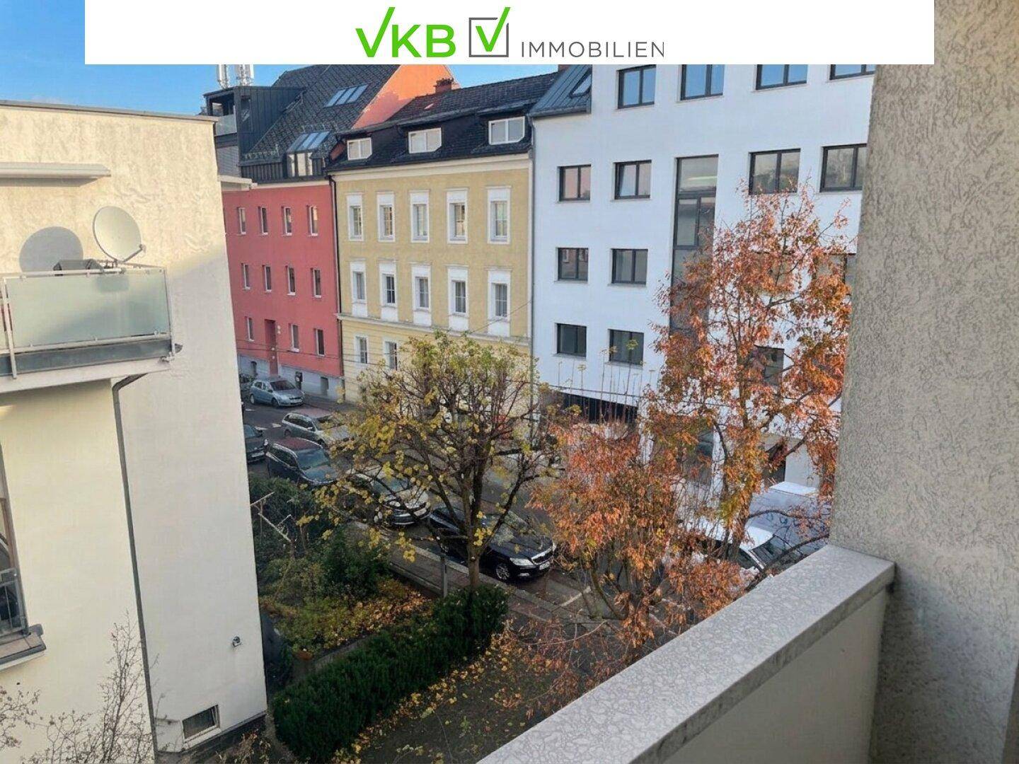 Balkon mit Blick auf die gegenüberliegenden Gebäude und die herbstliche Straße mit parkenden Autos.