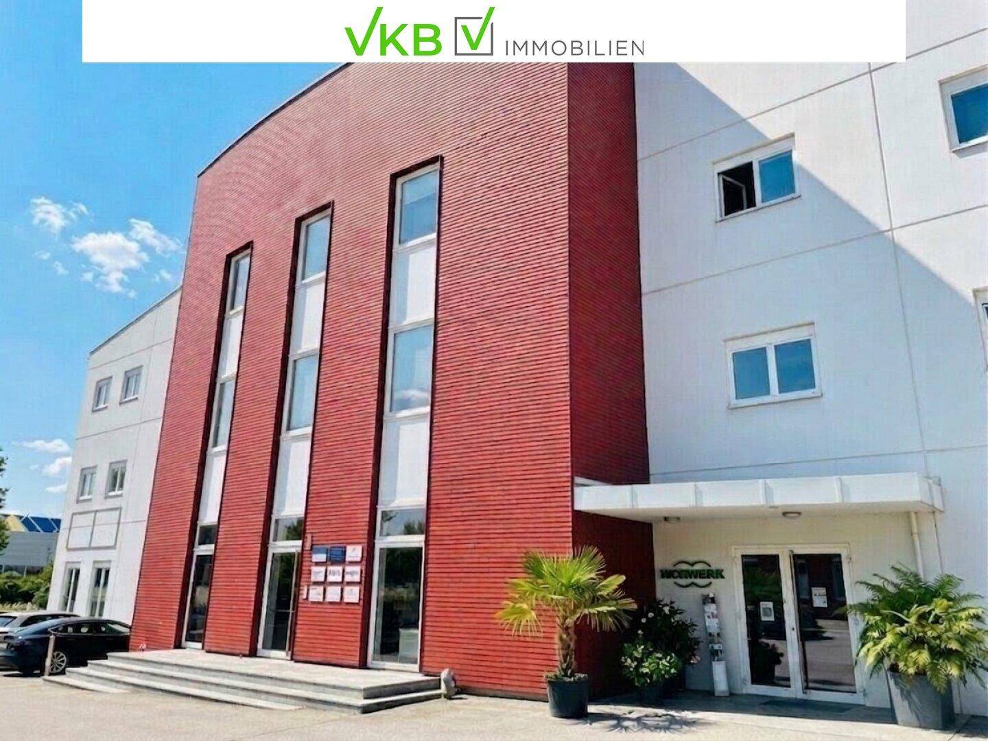 Eingangsbereich eines modernen Geschäftsgebäudes mit roter Fassade, hohen Fenstern und gepflegten Pflanzen.