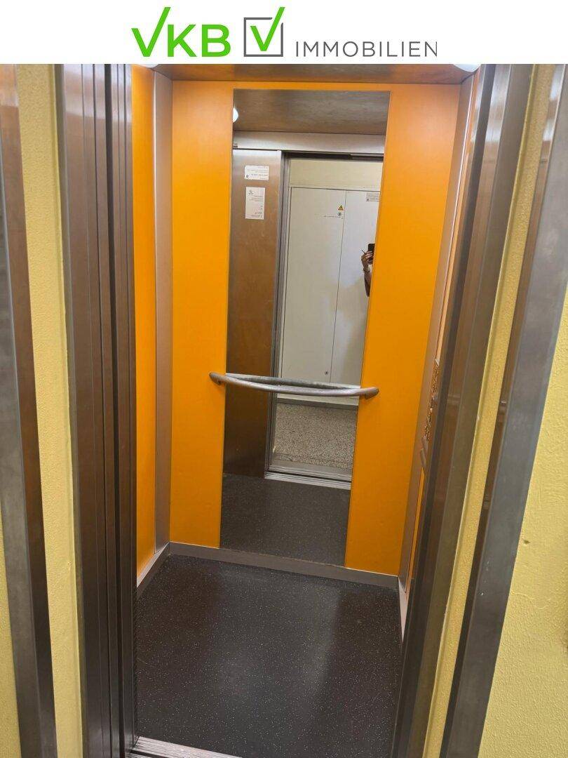 Offener Aufzug mit orangefarbenen Wänden und Metallhandlauf.