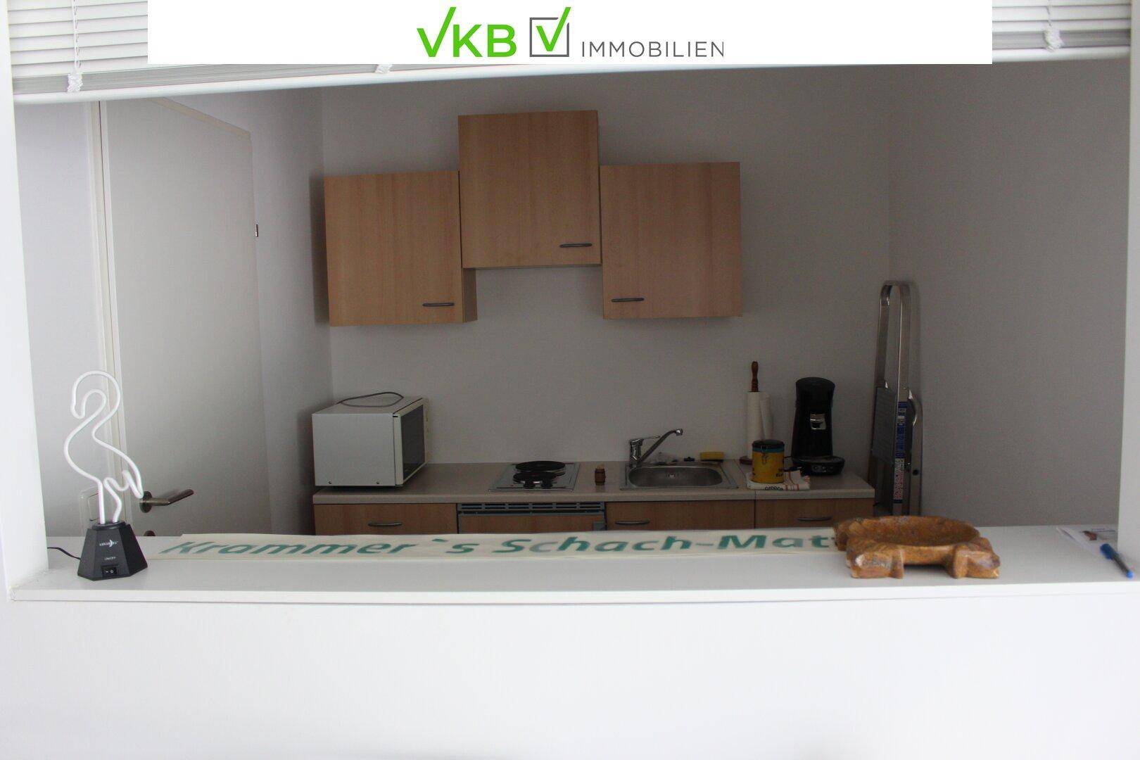 Kleine Küchenzeile mit Holzschränken, Spüle und Mikrowelle, sichtbar durch eine Wandöffnung.
