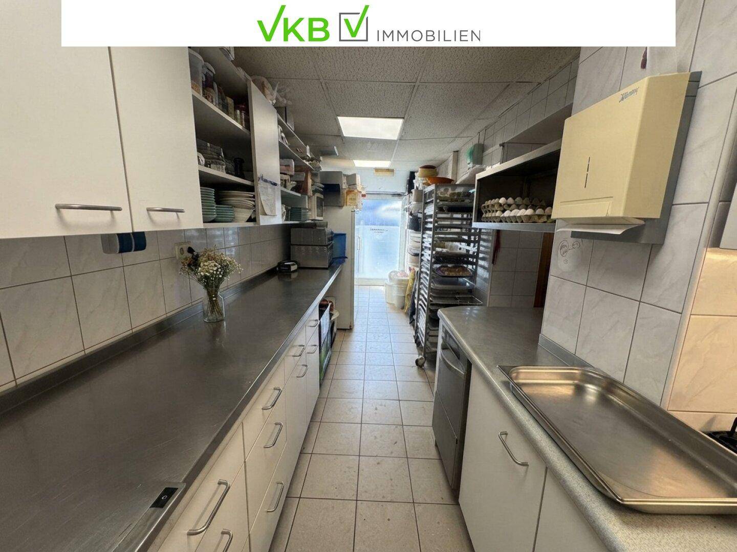 Professionelle Küche mit Edelstahl-Arbeitsflächen, weißen Hängeschränken und Fliesen an den Wänden.