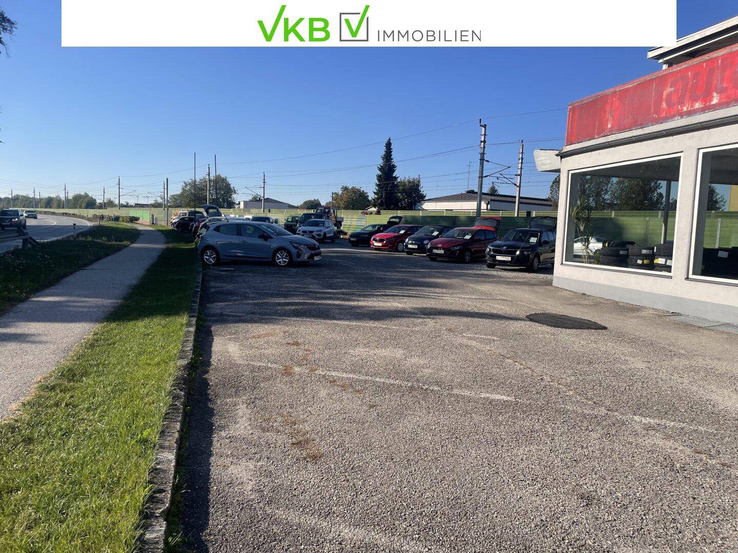 Blick auf den gepflasterten Außenparkplatz mit mehreren Fahrzeugen und angrenzendem Grünstreifen.
