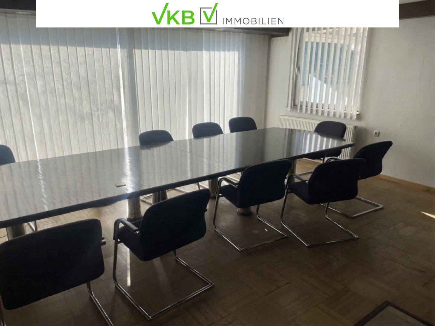 Konferenzraum mit langem Tisch und Stühlen, ausgestattet mit Jalousien an den Fenstern.