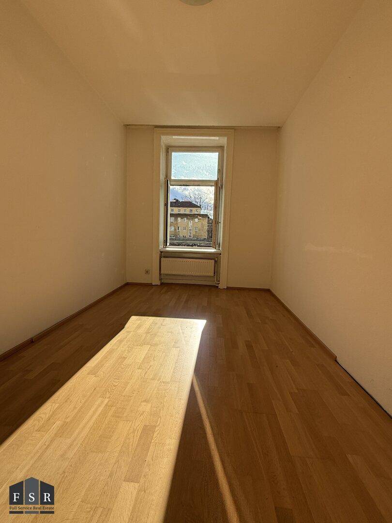 Langes, helles Zimmer mit Parkettboden und einem Fenster, das viel Tageslicht hereinlässt.