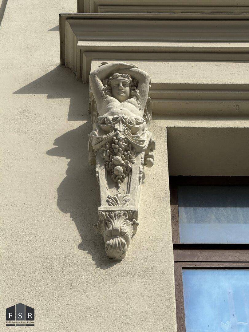 Nahaufnahme einer kunstvollen Fassadenverzierung mit figürlichen und floralen Elementen an einem historischen Gebäude.