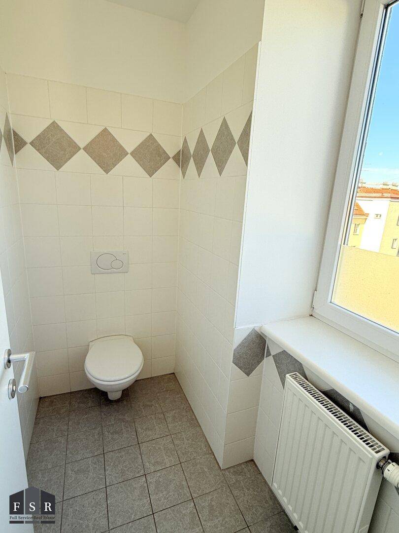 Separates WC mit Toilette, Fenster, Heizkörper und hellen Fliesen an Wänden und Boden.