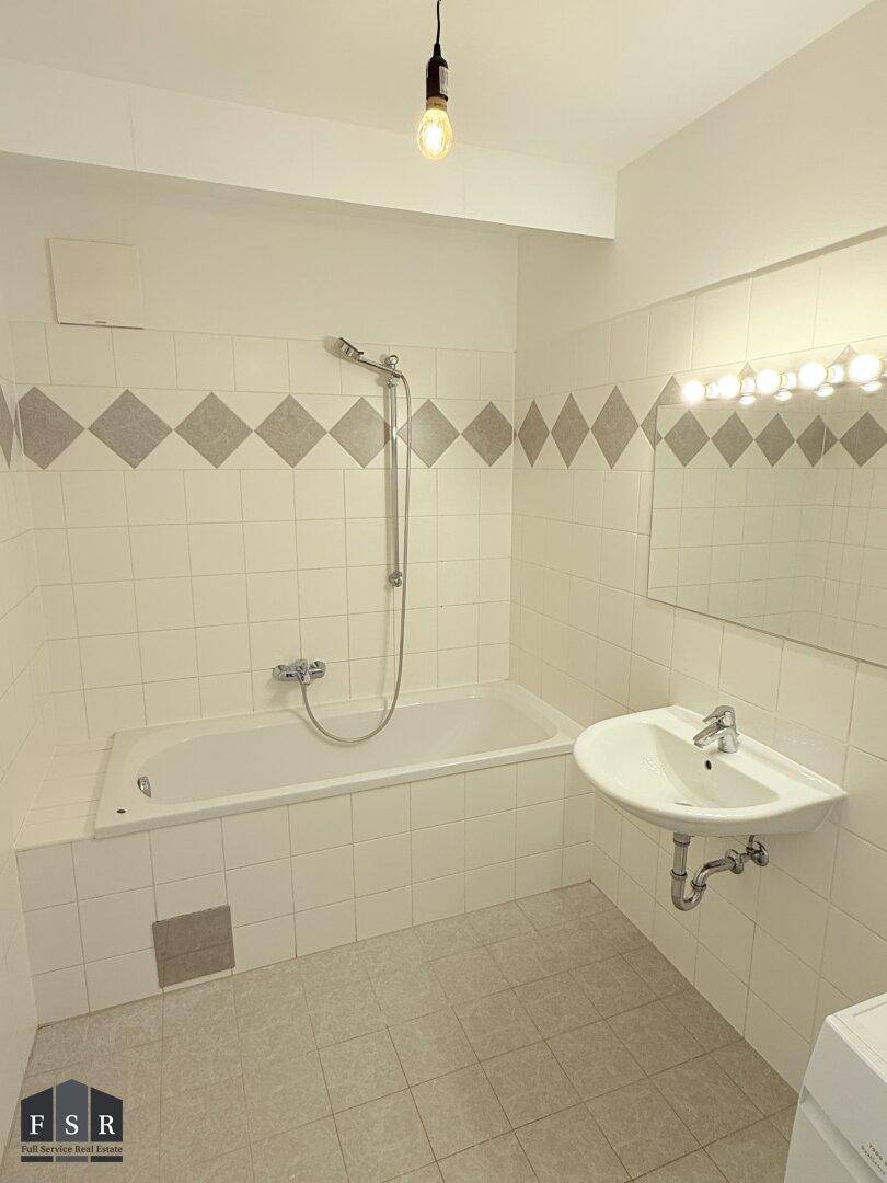 Badezimmer mit Badewanne, Waschbecken, Spiegel und hellen Fliesen an Wänden und Boden.