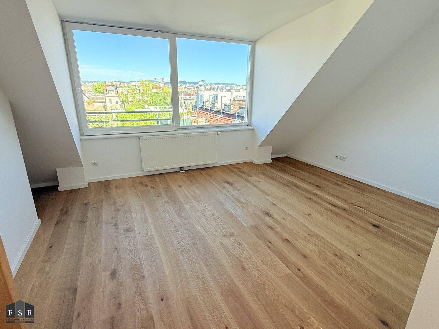 Helles Zimmer im Dachgeschoss mit Holzboden, großen Fenstern und weitem Blick über die Stadt.