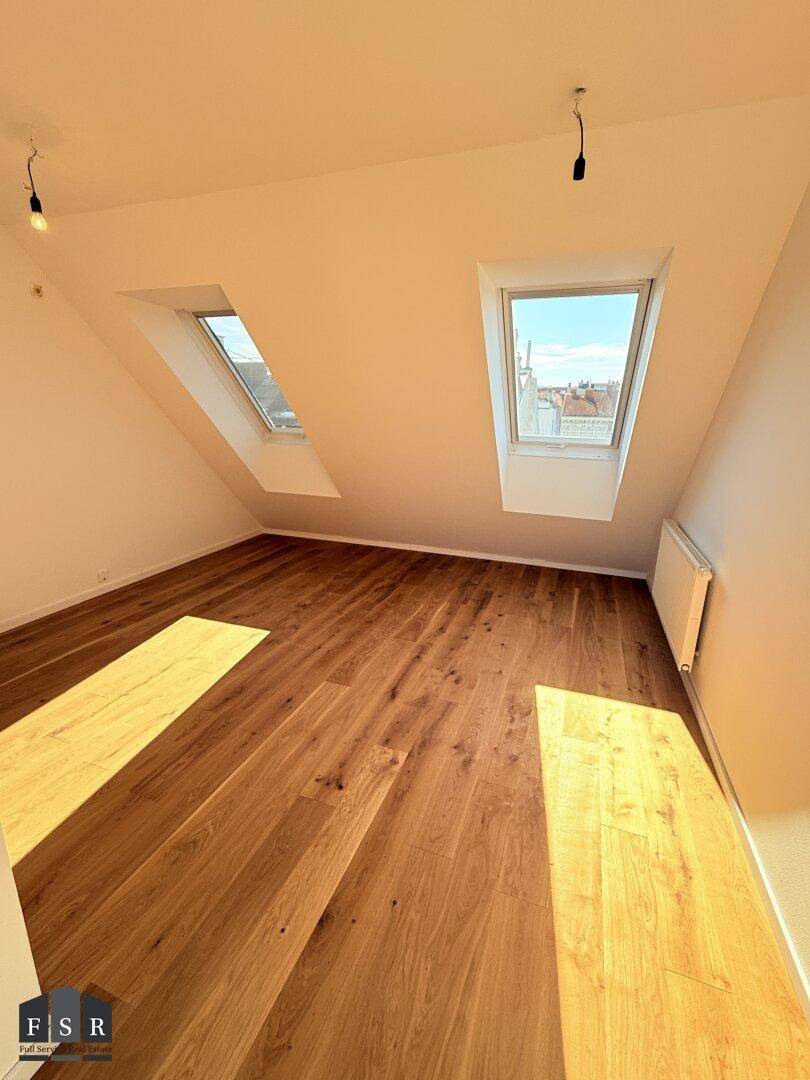 Sonniger Raum im Dachgeschoss mit Holzboden, zwei Dachfenstern und schrägen Wänden.