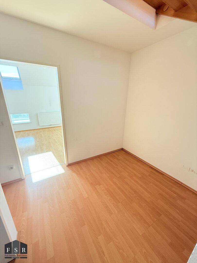 Leeres Zimmer mit Laminatboden, weiß gestrichenen Wänden und Zugang zu einem weiteren Raum.