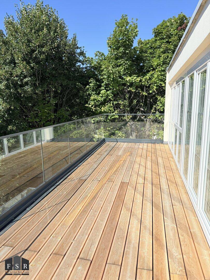 Sonniger Außenbereich mit Holzterrasse, Glasgeländer und umgebenden Bäumen.