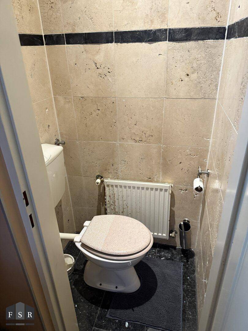Kleine separate Toilette mit beigen Fliesen, dunklem Boden und Heizkörper an der Wand.