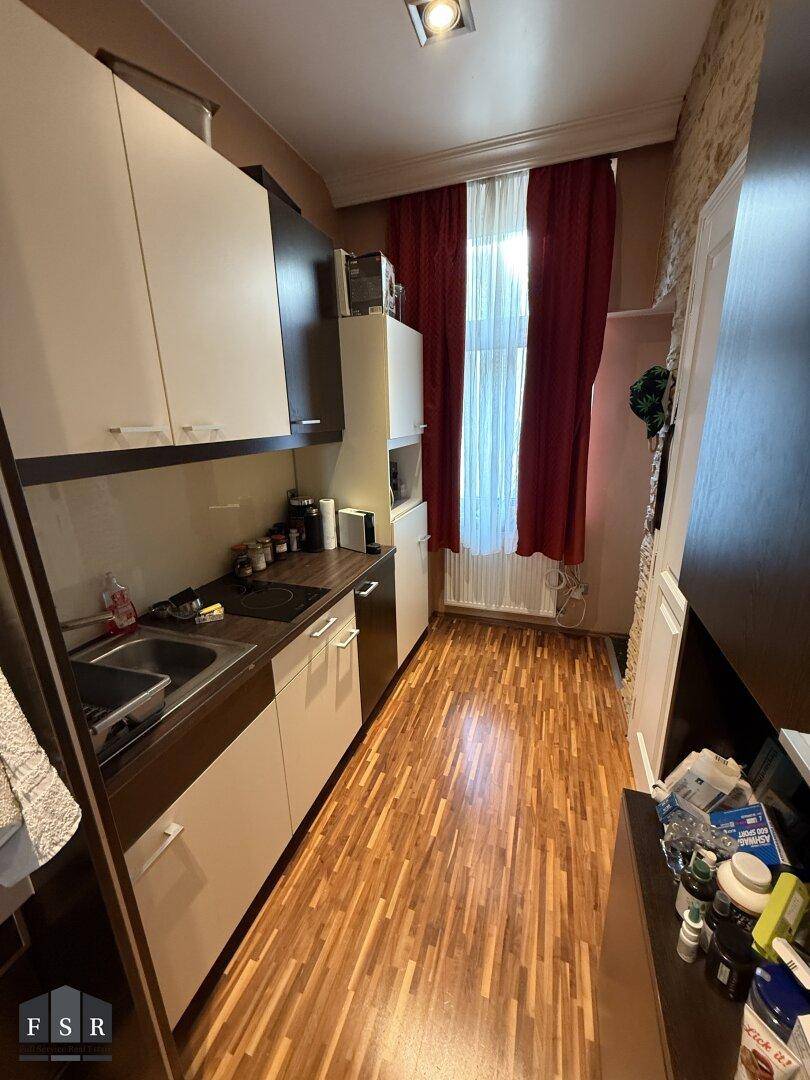 Schmale Küche mit weißen und dunklen Schränken, Holzboden und Fenster mit rotem Vorhang.