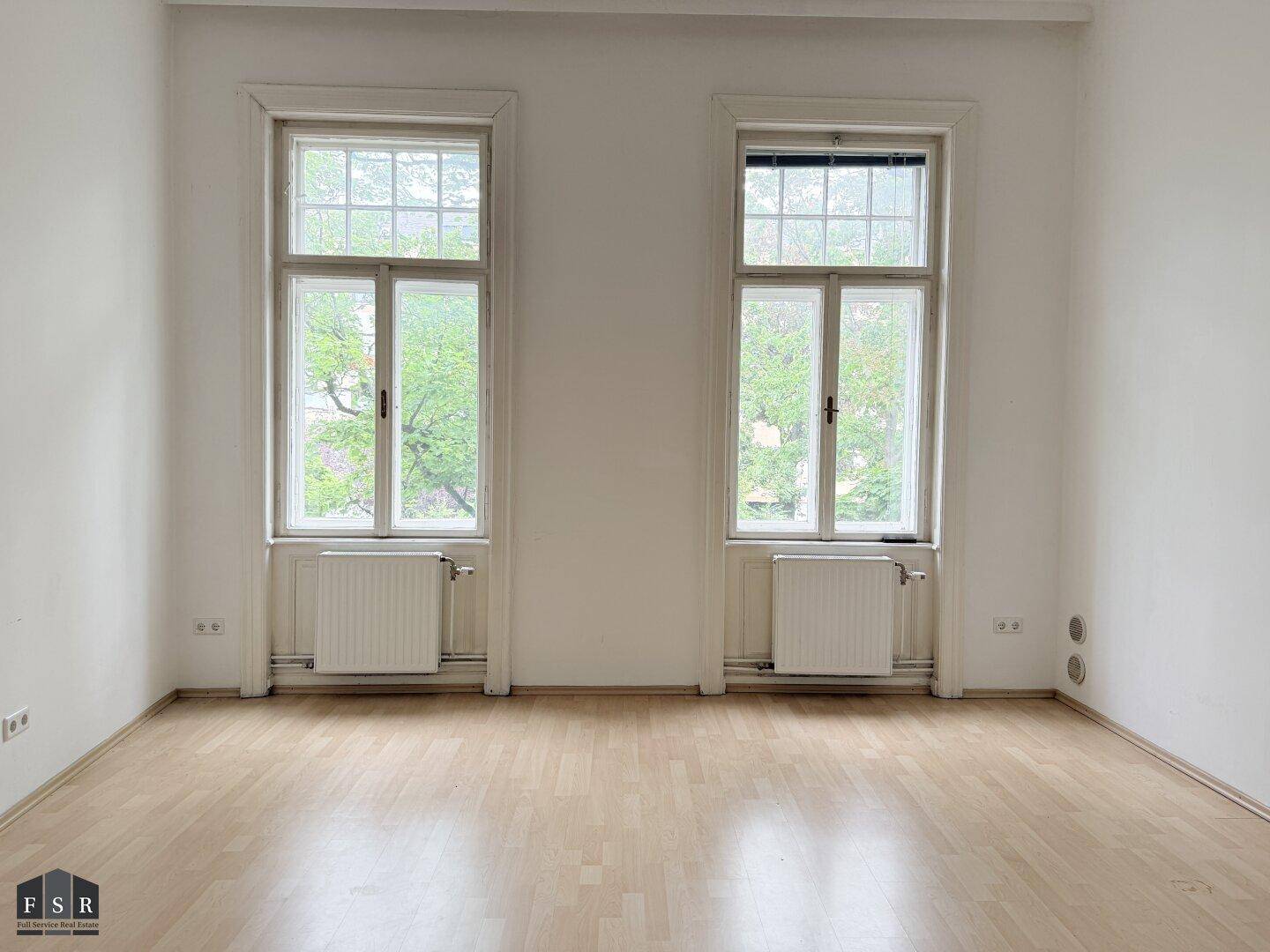 Helles Zimmer mit Laminatboden, zwei großen Fenstern und Blick ins Grüne.